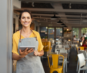 Business owner stands in front of her restaurant, holding a tablet and smiling at the camera.