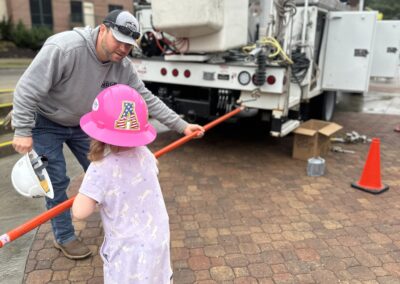 NRLP lineworkers helps a small child wearing a pink hard hat learn how to use linework gear near a bucket truck.