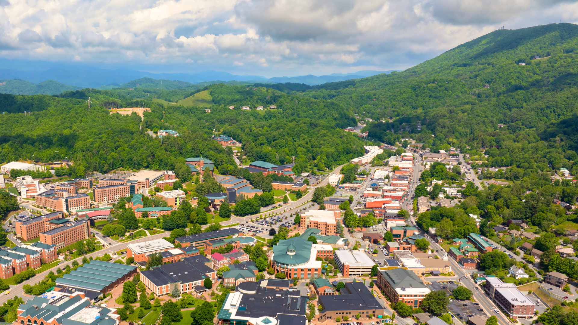 Aerial view of a portion of the town of Boone in the summer, with green-covered mountains in the background and blue skies above.