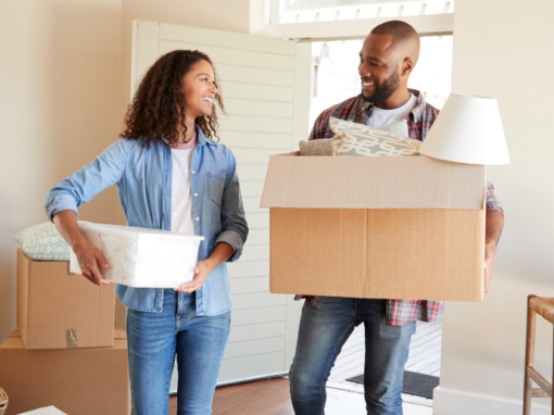A woman and man stand in a house with boxes packed, holding a box and a bin, smiling at each other.