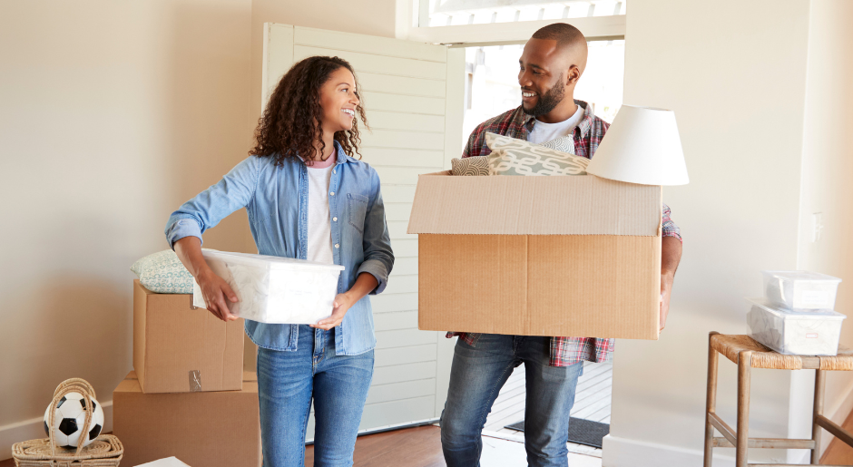 A woman and man stand in a house with boxes packed, holding a box and a bin, smiling at each other.