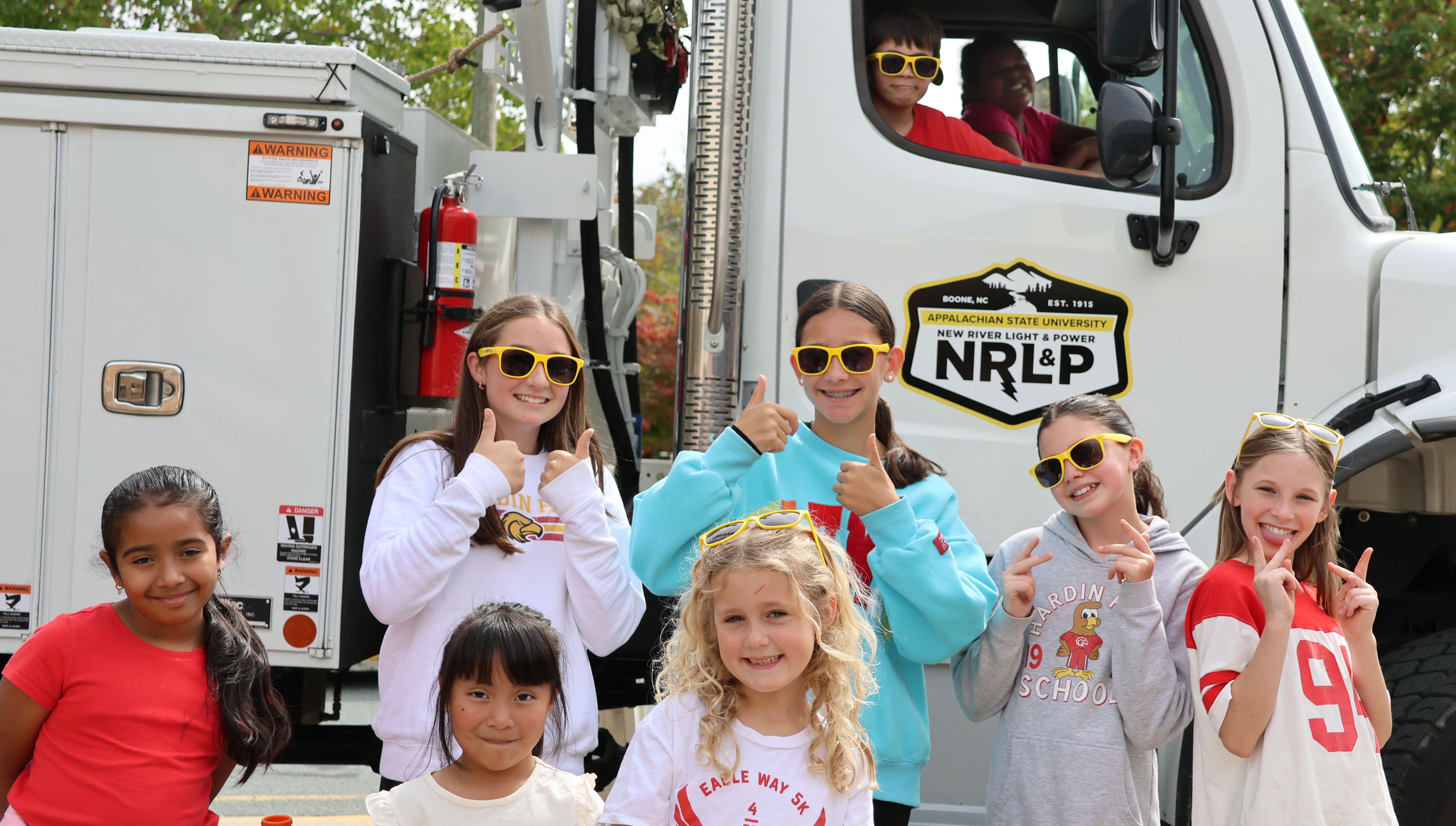 A group of students stand in front of and in a bucket truck, smiling and giving thumbs up